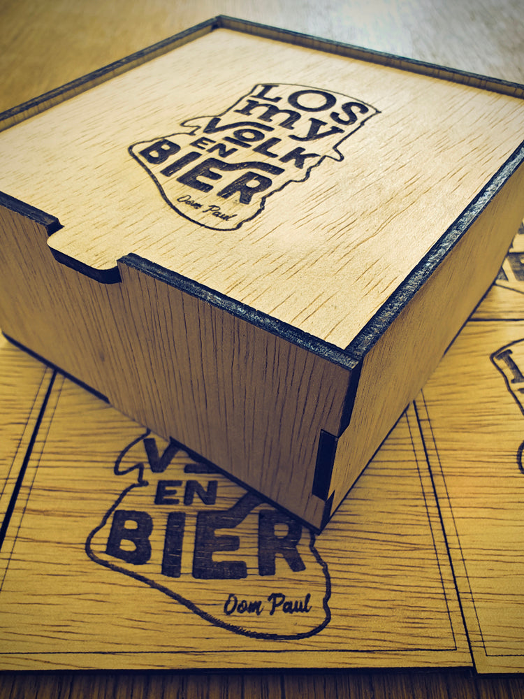A laser-engraved wooden coaster with the text 'Los my Volk en Bier' Oom Paul' on it, displayed inside a wooden storage box.